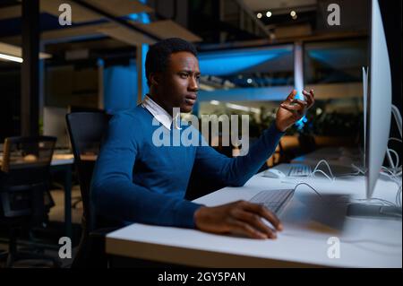 L'uomo in occhiali lavora al computer, stile di vita dell'ufficio. Uomo al tavolo, interni scuri, ambiente di lavoro moderno Foto Stock