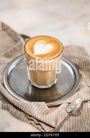 Bicchiere di caffè bianco piatto appena preparato con latte in schiuma a forma di cuore su vassoio argento su lino beige. Bevanda idilliaca per la colazione Foto Stock