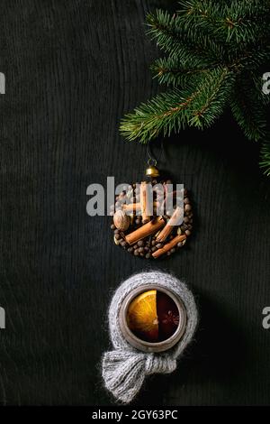 VIN brulé rosso caldo in tazza di ceramica in sciarpa con spezie a forma di palla di Natale su abete, fetta di arancia su sfondo nero di legno. Bevande per le vacanze Foto Stock