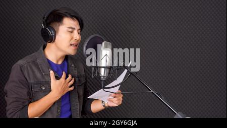 Giovane asiatico con le cuffie cantano mentre tiene il testo in mano davanti alle pareti nere dell'insonorizzazione. Musicisti che producono musica professionale Foto Stock