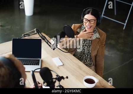 vista ad alto angolo del gioioso host radio asiatico in occhiali e cuffie ridendo mentre si guarda un collega sfocato Foto Stock