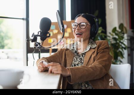 la radio asiatica ha un'accoglienza positiva in occhiali e cuffie che tengono lo smartphone mentre ride durante il podcast Foto Stock