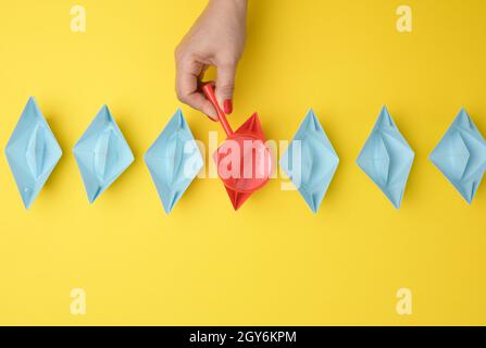 la mano femmina tiene una lente d'ingrandimento su una fila di barche di carta su uno sfondo giallo. Concetto di ricerca di talenti, reclutamento, interviste di reclutamento Foto Stock