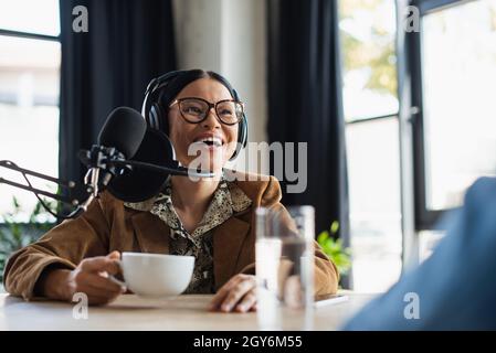 felice radio asiatica ospite in occhiali e cuffie ridendo mentre tiene la tazza in studio di trasmissione Foto Stock