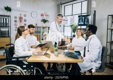 Il medico caucasico bearded scuotendo le mani ed il saluto i suoi colleghi multietnici durante il seminario medico all'ufficio. Utente su sedia a rotelle femminile che lavora con i colleghi su un progetto comune. Foto Stock