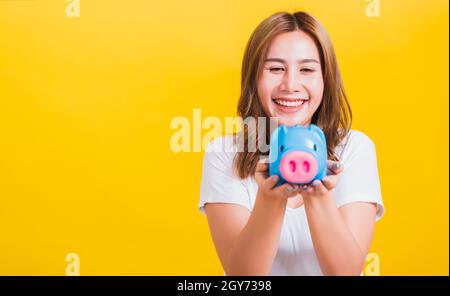 Ritratto Asian Thai bella giovane donna felice sorridente tenendo blu piggy banca con un sacco di soldi e guardando a piggybank, studio girato isolato su Foto Stock