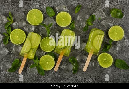 Primo piano di diversi papsicli surgelati di succo di frutta con fette di lime fresche, foglie di menta verde e cubetti di ghiaccio sulla superficie grigia del tavolo, vista dall'alto, diretta Foto Stock