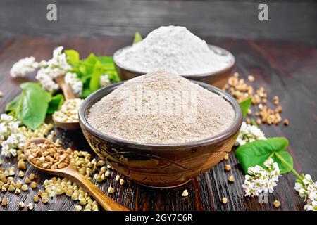 Farina di grano saraceno di cereali marroni e verdi in due ciotole, cucchiai con diverse semole di grano saraceno, fiori e foglie sullo sfondo di un bo di legno Foto Stock