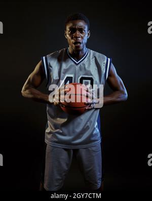 Aggressivo giocatore di basket pone con palla in studio, sfondo nero. Baller professionista in abbigliamento sportivo, sportivo alto Foto Stock