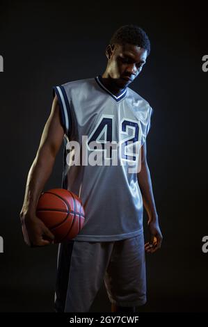 Aggressivo giocatore di basket pone con palla in studio, sfondo nero. Baller professionista in abbigliamento sportivo, sportivo alto Foto Stock