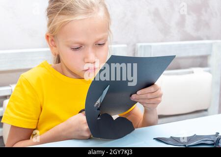 Una ragazza taglia una decorazione di Halloween a casa da carta nera. Fai da te artigianato decorazioni per la casa. Il bambino fa i mestieri di carta, origami Foto Stock