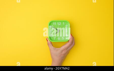 la mano della donna tiene una sveglia verde quadrata, l'ora è dieci - otto alla mattina. Concetto di Early Rise, sfondo giallo Foto Stock