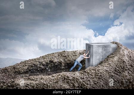 l'uomo spinge un enorme cubo di cemento strisciando nel terreno. concetto di determinazione e forza. Foto Stock