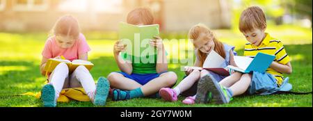 Bambini con zaini seduti nel parco sull'erba vicino alla scuola. Alunni con libri e zaini all'aperto. Foto Stock