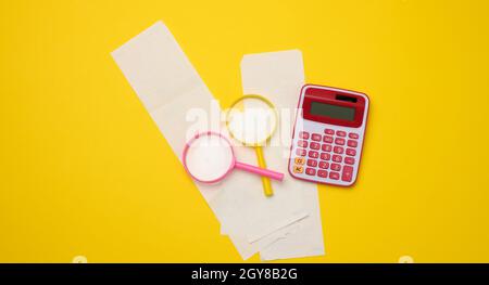 una pila di assegni di carta, una calcolatrice di plastica rosa e una lente di ingrandimento su sfondo giallo. Concetto di audit del budget familiare, ricerca di risparmi Foto Stock