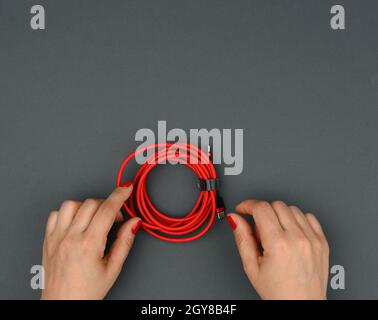 la mano femmina tiene il cavo ritorto per caricare i dispositivi mobili in un involucro di tessuto rosso su uno sfondo nero, primo piano Foto Stock