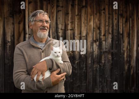 Un anziano uomo detiene un carino gattino bianco e sorrisi alla telecamera, messa a fuoco selettiva, con abbondanza di spazio di copia Foto Stock
