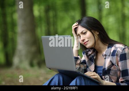 Gara mista triste donna che controlla il portatile seduto in una foresta Foto Stock