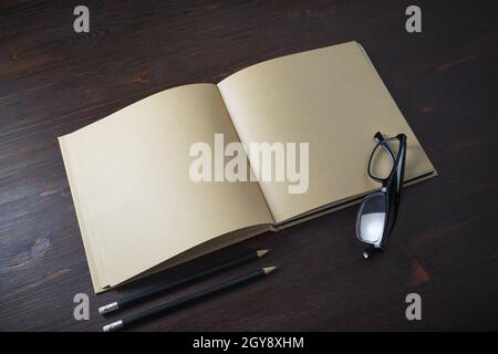 Libro aperto con pagine bianche di carta artigianale, matite e bicchieri su sfondo di legno. Foto Stock