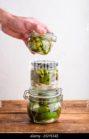 Mano maschile tenendo uno dei tre vasi di vetro di peperoni Jalapeno appena decapati con erbe e aglio. Copia spazio Foto Stock