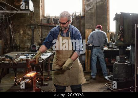 Maturo barbuto fabbro in grembiule lavorando con il ferro in officina con il suo collega in background Foto Stock