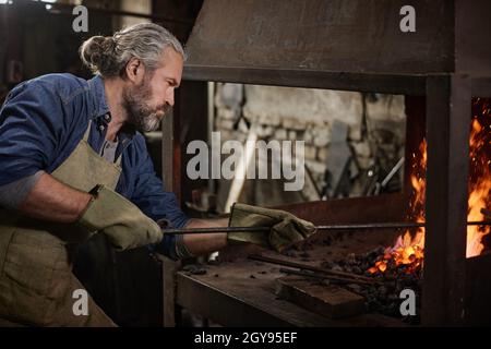 Lavoratore maturo bearded che mette i carboni nella fornace durante il suo lavoro nel negozio di fabbro Foto Stock