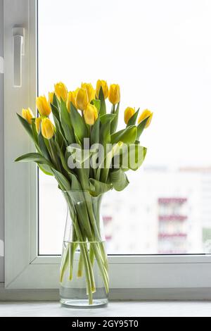 fiori di tulipano gialli freschi in vaso di vetro sul davanzale della finestra a casa con paesaggio urbano sullo sfondo Foto Stock