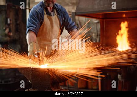 Fabbro in grembiule forgiando il ferro con il martello durante il lavoro in officina Foto Stock