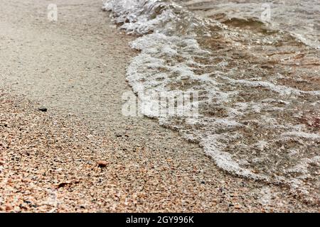 Una piccola onda di mare con schiuma. Schiuma di mare sulla spiaggia di sabbia con acqua limpida. Spiaggia di sabbia e schiuma di mare Macro con fuoco stretto sfondo. Sfondo estivo. Foto Stock