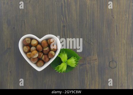 Nocciole fresche non sgusciate in un piatto a forma di cuore con foglie verdi in un piano in alto stendere la vita su un tavolo di legno con copyspace Foto Stock