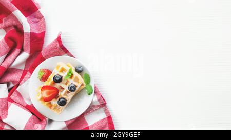 Piatto bianco su asciugamano rosso da cucina con cialde fatte in casa, mirtillo, tagli di fragole, foglia di menta, polvere di zucchero su sfondo grigio Foto Stock