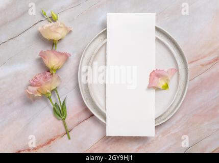 Menù di nozze adagiato su una piastra in ceramica su un tavolo in marmo decorato con fiori e nastri. Scena mock-up con biglietti d'auguri di carta vuoti. Femminile f Foto Stock