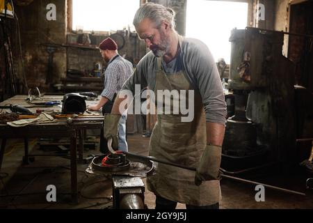 Maturo barbuto fabbro che fa il dettaglio dal ferro insieme con collega in background nel negozio di fabbro Foto Stock