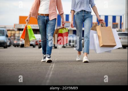 Uomo e donna con borse di cartone sul parcheggio del supermercato. Clienti felici che trasportano acquisti dal centro commerciale, veicoli in background Foto Stock