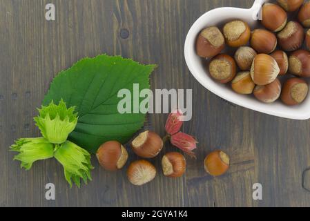 Nocciole fresche non sgusciate in un piatto a forma di cuore con foglie verdi in un piano in alto stendere la vita su un tavolo di legno con copyspace Foto Stock