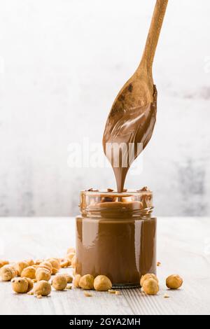 Crema di cioccolato in vaso di vetro con nocciole e cucchiaino gocciolante, su tavolo di legno Foto Stock