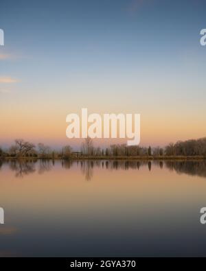 Una casa, gli alberi e il tramonto creano un riflesso su un lago fermo Foto Stock