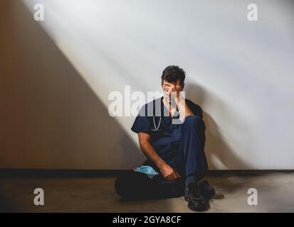 Medico esausto che indossa scrub seduti sul pavimento in un cerotto di luce. Foto Stock