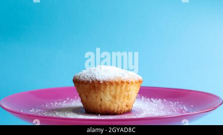 Una cagliata cosparso di zucchero a velo su un piatto rosa, su sfondo blu, vista laterale. Dessert, un piccolo cupcake. Concetto di cibo. Biscotti bianchi cotti Foto Stock