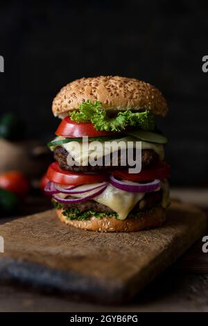 Hamburger con pomodori, cipolle, cetrioli, lattuga e formaggio fuso servito su un rustico tavolo di legno con patatine fritte e verdure in background Foto Stock