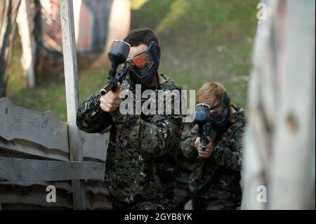 Due giocatori di paintball si imitano con le pistole dal rifugio sul parco giochi. Sport estremo con arma pneumatica e pallottole o marcatori di vernice, gioco militare Foto Stock