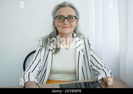 Donna d'affari grigia senior con capelli in giacca a righe con occhiali sta lavorando nel suo ufficio seduto alla scrivania. Buon pensionamento, occupazione e. Foto Stock