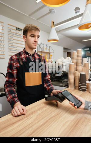 il barista sorridente accetta il pagamento dal telefono cellulare tramite la macchina pos nel bar. il cliente che tiene il telefono vicino al terminale nfc effettua il pagamento mobile senza contatto. Foto Stock