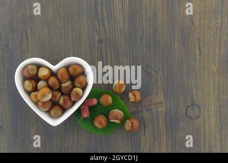 Nocciole fresche non sgusciate in un piatto a forma di cuore con foglie verdi in un piano in alto stendere la vita su un tavolo di legno con copyspace Foto Stock