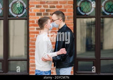 Giovane coppia gay che indossa una maschera medica, abbracciando e baciando in città. Foto Stock