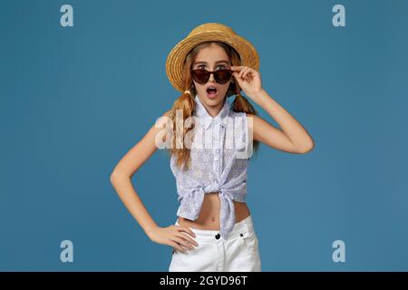 sorpresi bambina in estate cappello e occhiali da sole vi guarda su sfondo blu. vendita estate Foto Stock