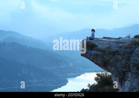 Ritratto a corpo pieno di una donna che fa esercizio di yoga in cima a una scogliera in un ambiente blu Foto Stock