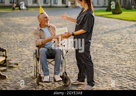 Gioioso uomo anziano, ricoverando il paziente in sedia a rotelle indossando il cappello di compleanno ricevere il regalo e le congratulazioni da giovane infermiera Foto Stock
