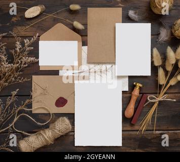 Cancelleria per matrimoni allestita con busta posta su un tavolo di legno con decorazione bohémien intorno. Scena mock-up con biglietti d'auguri in carta bianca e asciugati Foto Stock