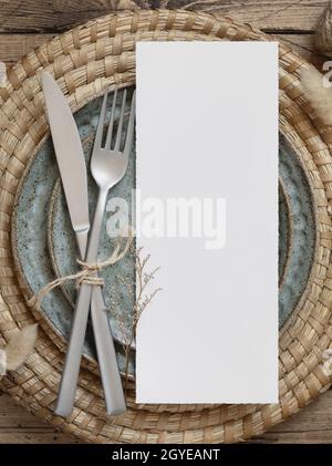 Carta da menu vuota su piastra con forchetta e coltello su tavola in legno con decorazioni bohémien e piante essiccate, vista dall'alto. Boho mockup carta da matrimonio Foto Stock